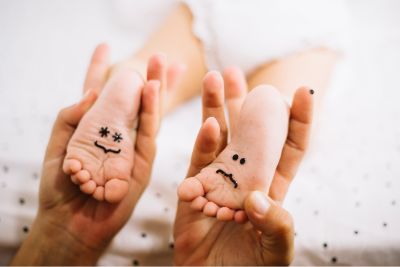 personne qui tient les pieds d'un bébé, sur lesquels sont dessiné un visage avec le sourire, dans chacune de ses mains